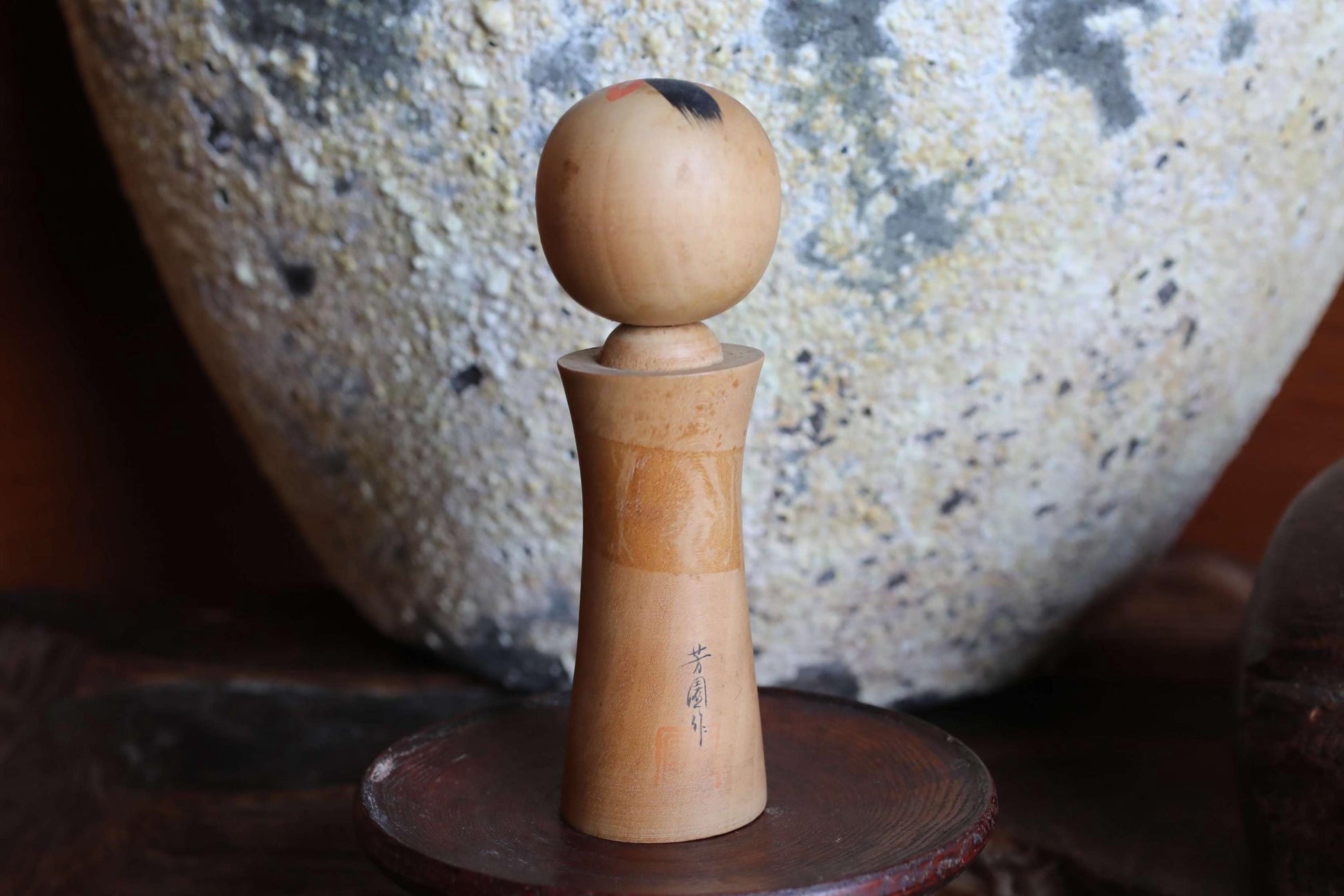 A wooden object with a round top and a textured body, placed on a wooden stand in front of a large, textured stone or ceramic piece.