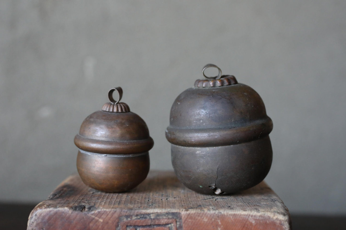 Two antique brass containers with round tops sit on a wooden surface against a plain background.