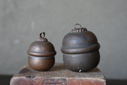 Two antique brass containers with round tops sit on a wooden surface against a plain background.