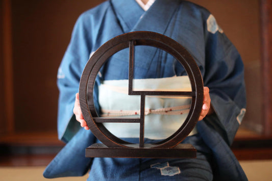 A person wearing a blue kimono holds a circular wooden object with a geometric design in front of them.