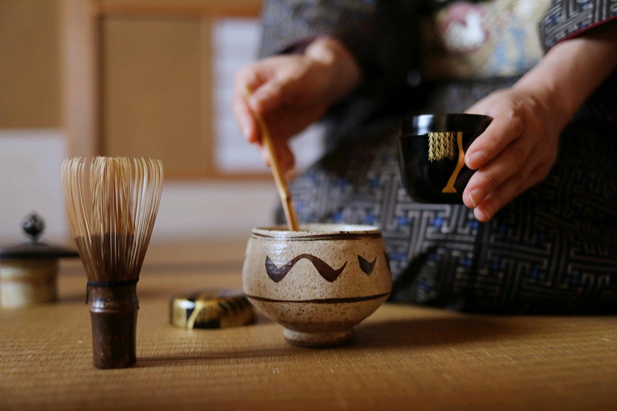 A person is seated at a table, preparing to make matcha tea. They are holding a black cup with a gold design, and a small ceramic bowl with a brown and black design. The table also has a small ceramic whisk and a small ceramic cup.
