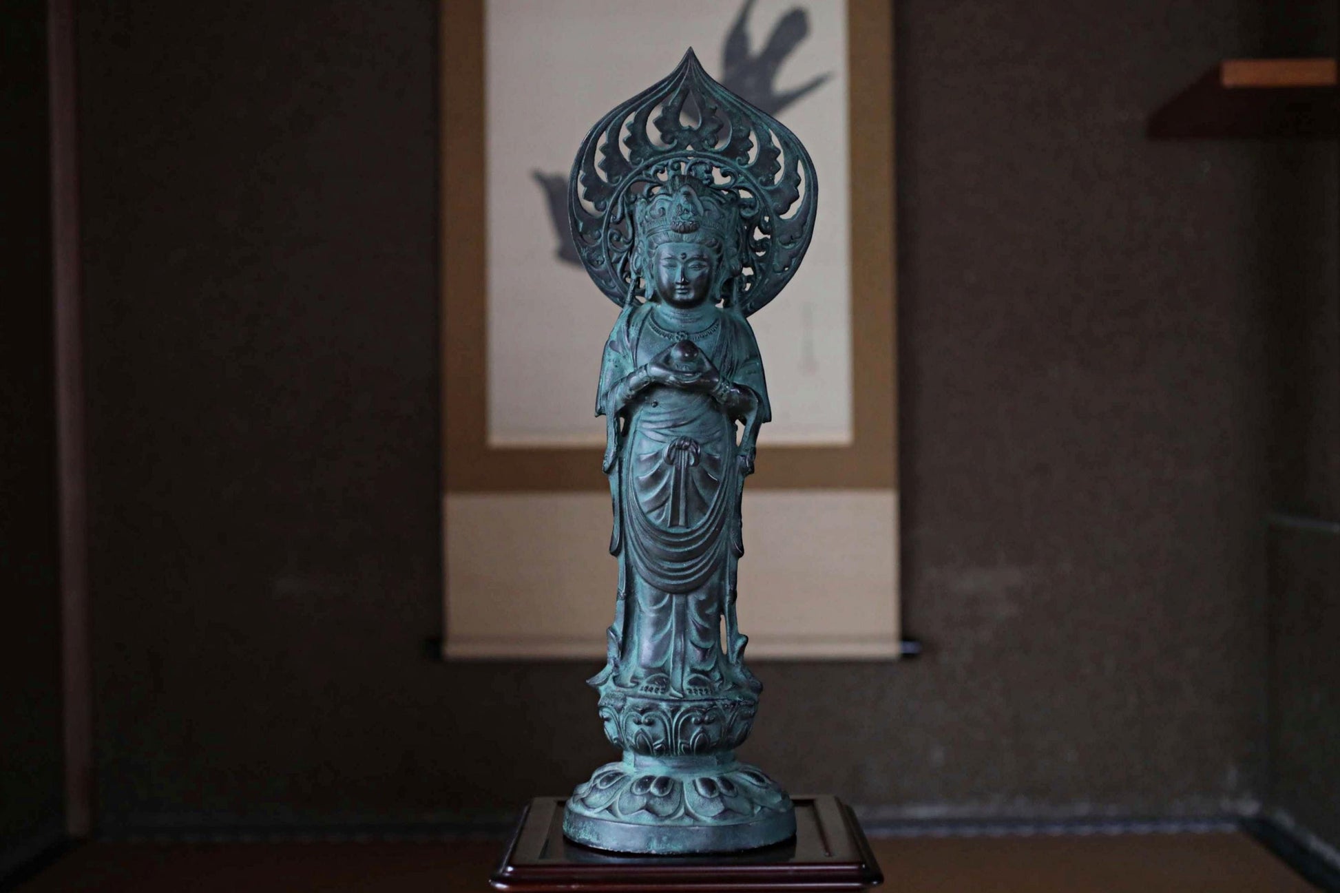 A bronze statue of a Buddhist deity, likely a Buddha, is displayed on a wooden stand in a dimly lit room.