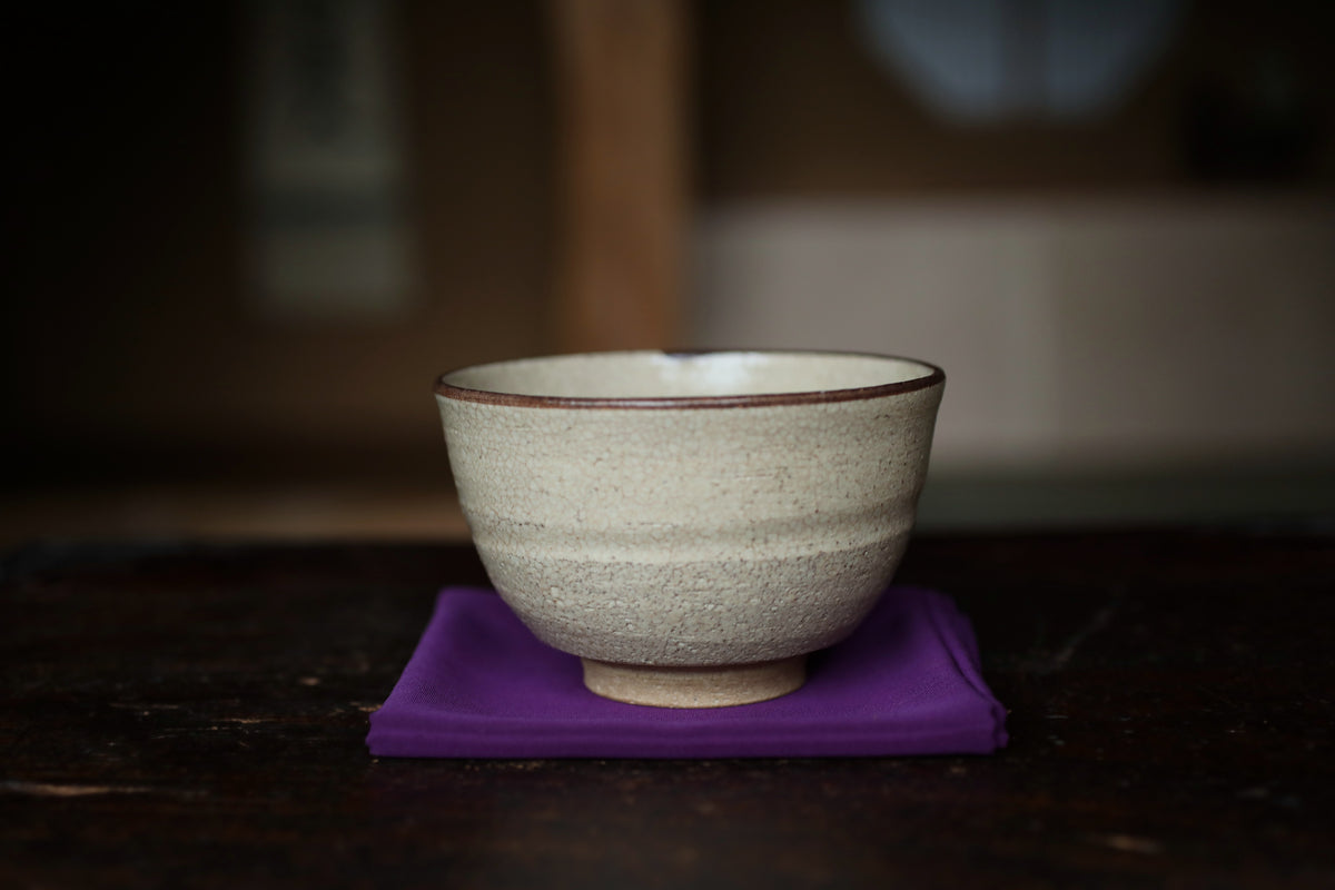 A white ceramic bowl with a brown rim sits on a purple cloth on a wooden surface.