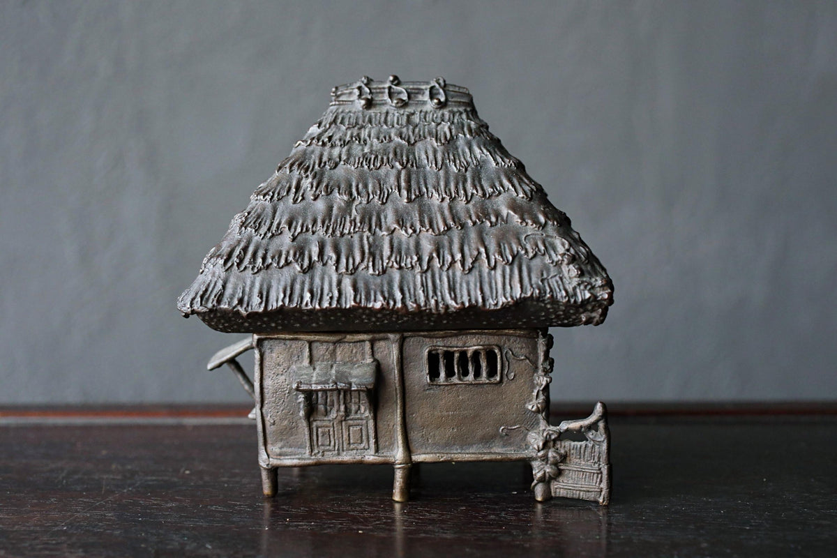 A small, intricately designed metal model of a house with a thatched roof, sitting on a dark surface.