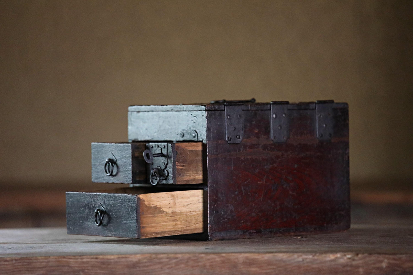 An old, wooden chest with metal clasps and drawers sits on a wooden surface, its weathered appearance suggesting it has been well-used over time.
