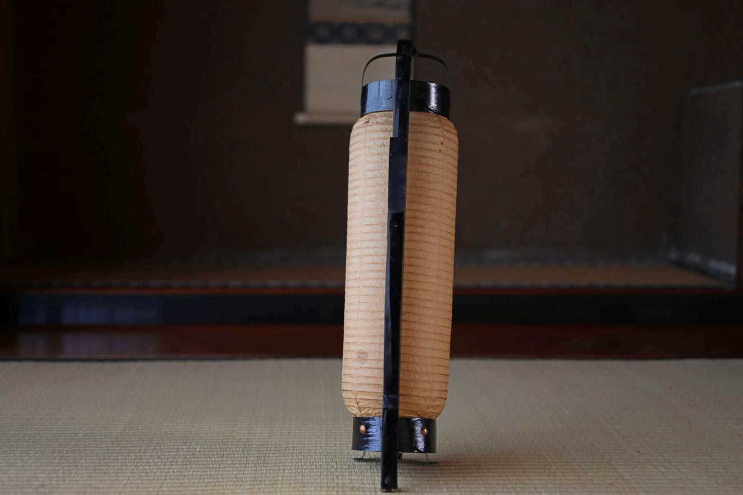 A traditional Japanese paper lantern with a black handle stands on a tatami mat, creating a serene and minimalist atmosphere.