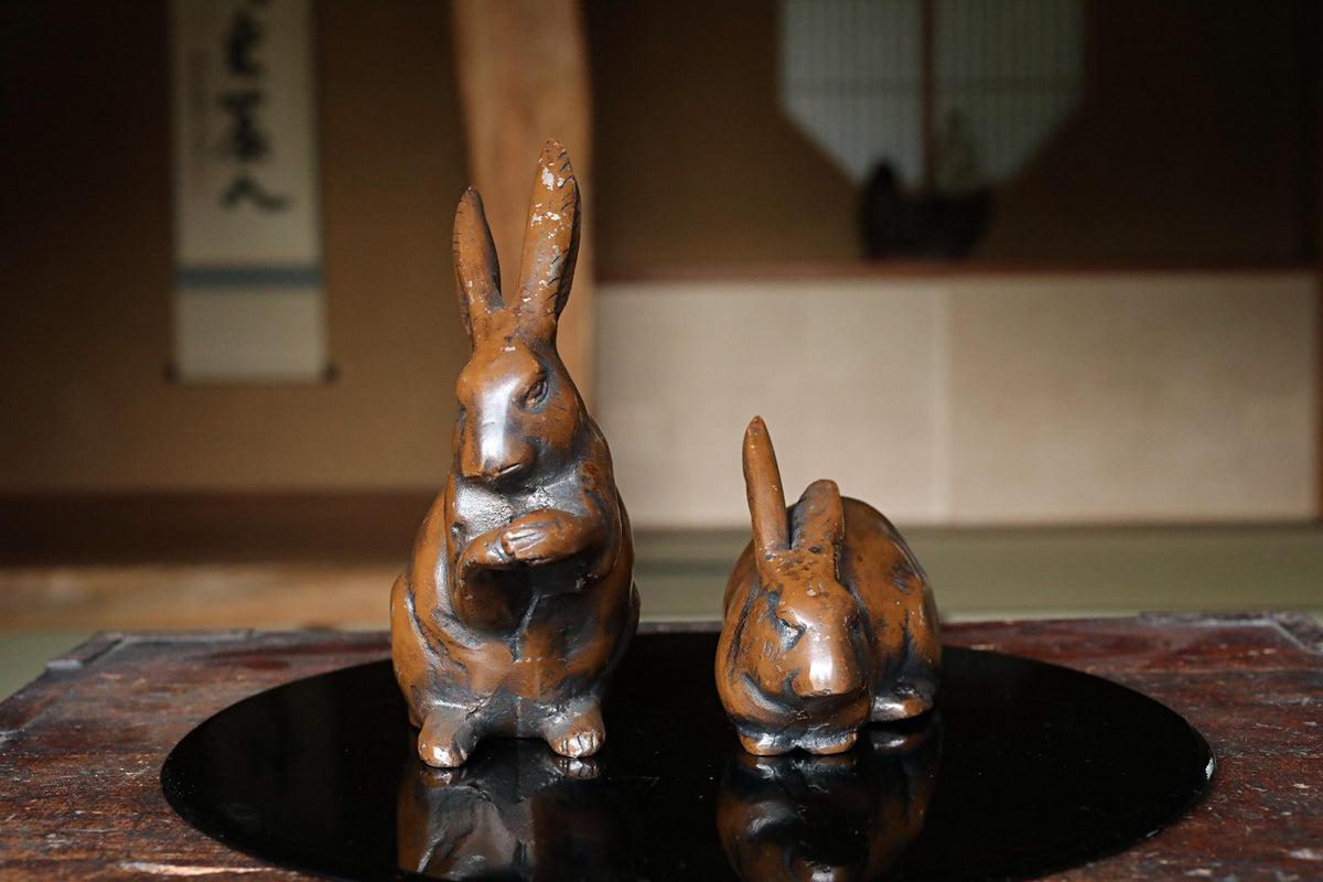 Two bronze rabbit figurines are displayed on a black circular surface, with a blurred background suggesting an indoor setting.