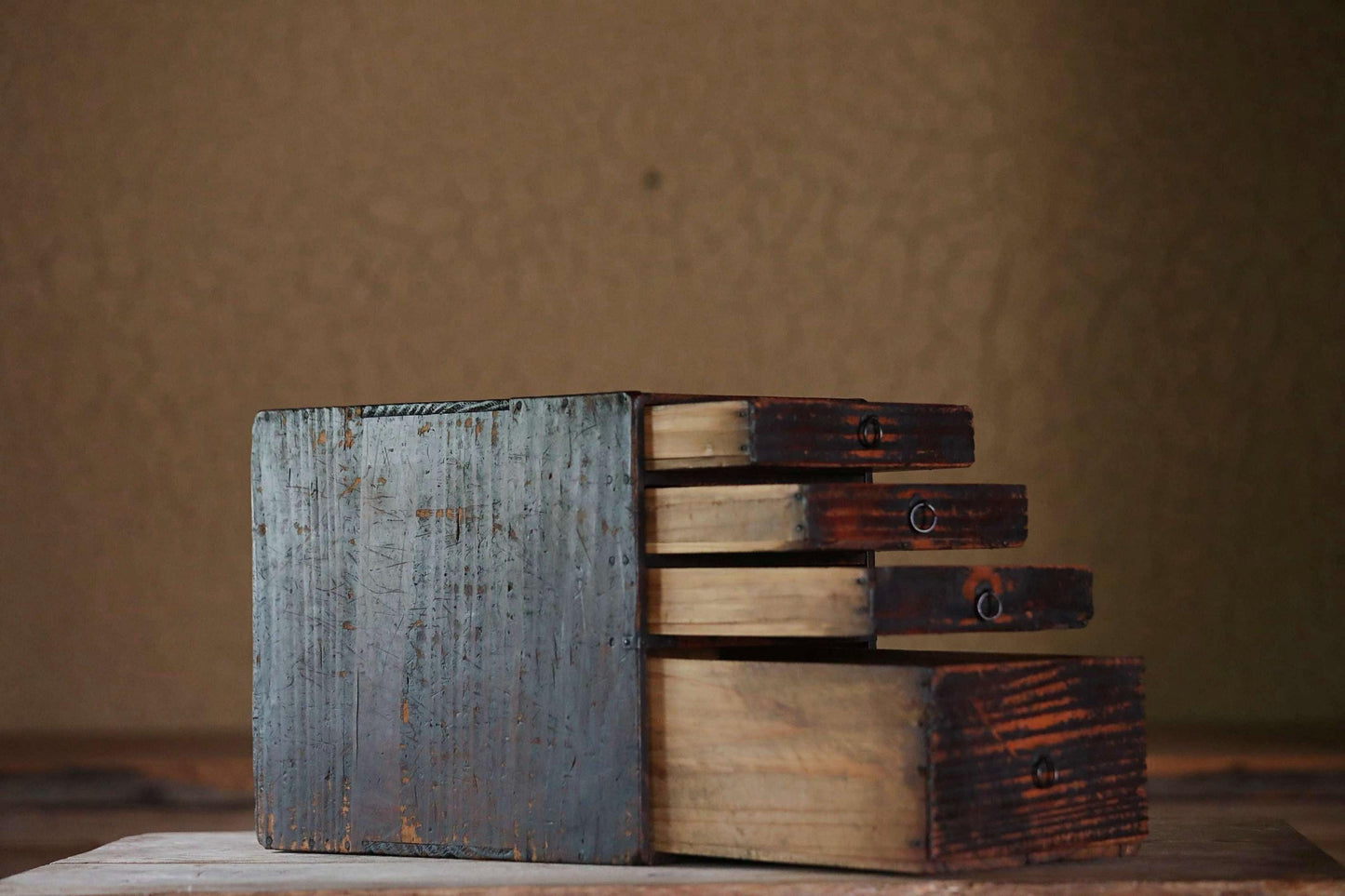 An old, weathered wooden drawer with four open compartments sits on a wooden surface against a textured wall.