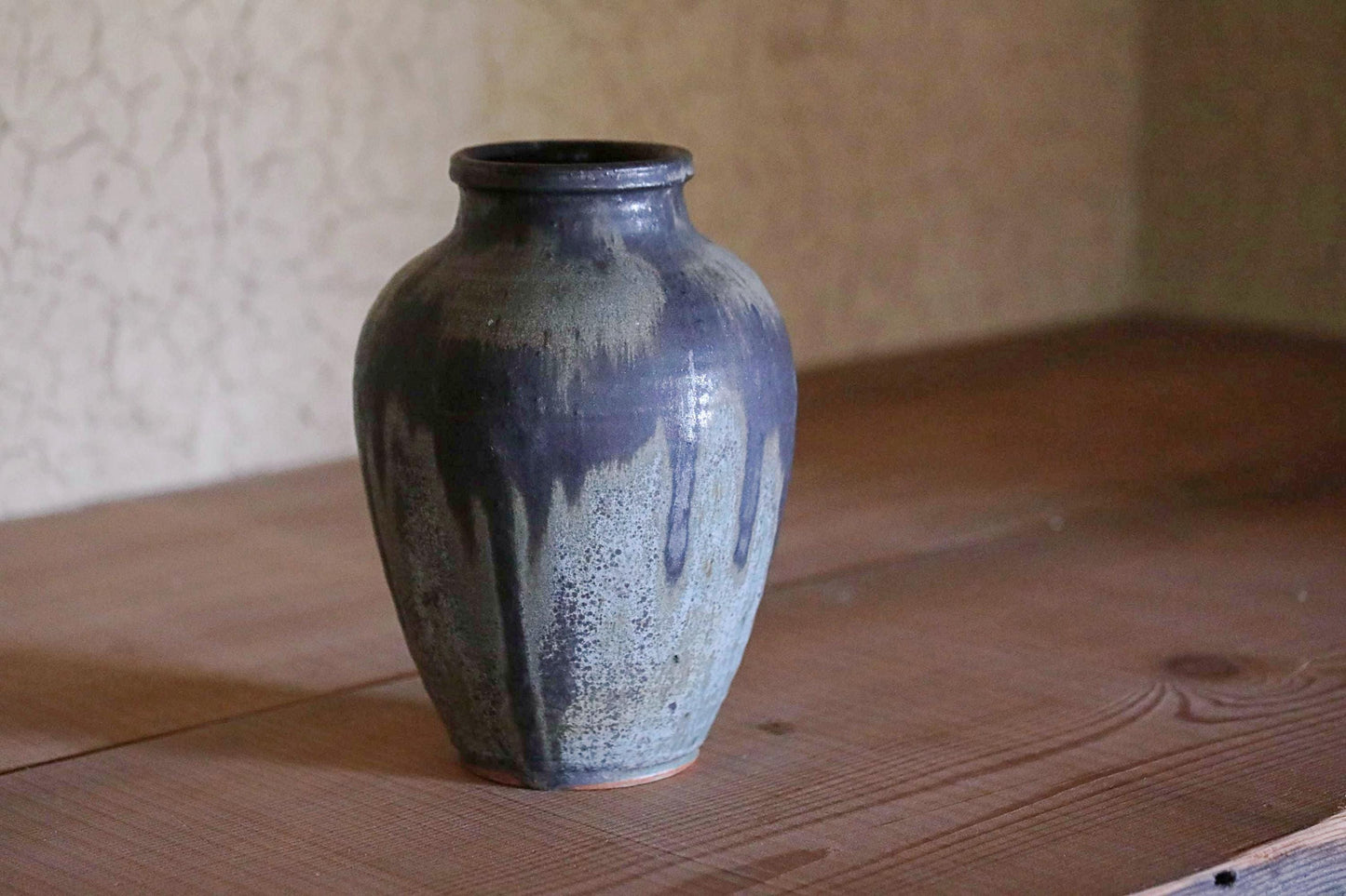 A dark blue ceramic vase with a textured surface sits on a wooden surface against a plain wall.
