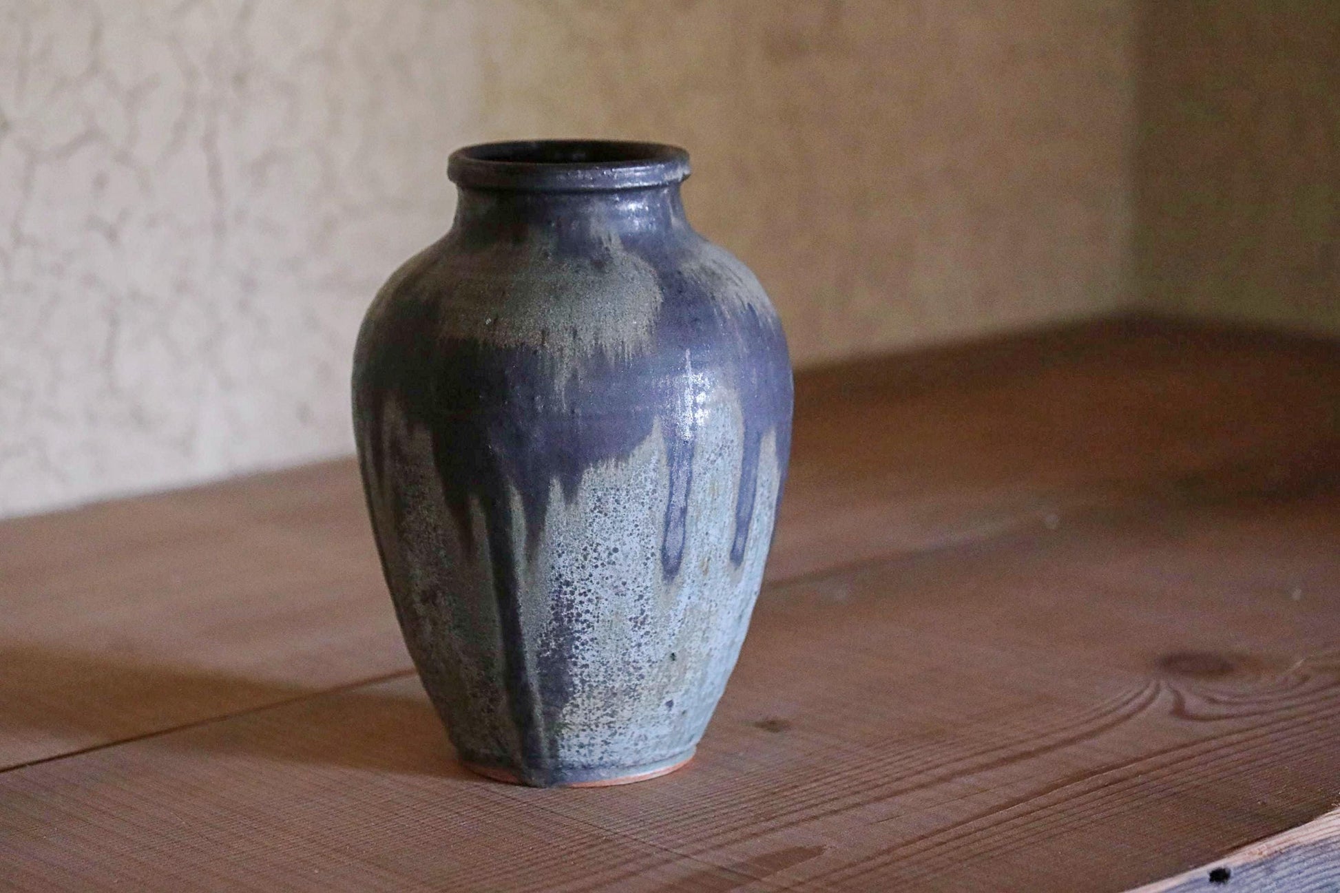 A dark blue ceramic vase with a textured surface sits on a wooden surface against a plain wall.