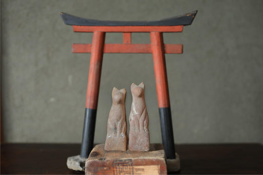 two small statues of cats, one on each side of a red and black torii gate, which is a traditional Japanese gate used at the entrance to a Shinto shrine.