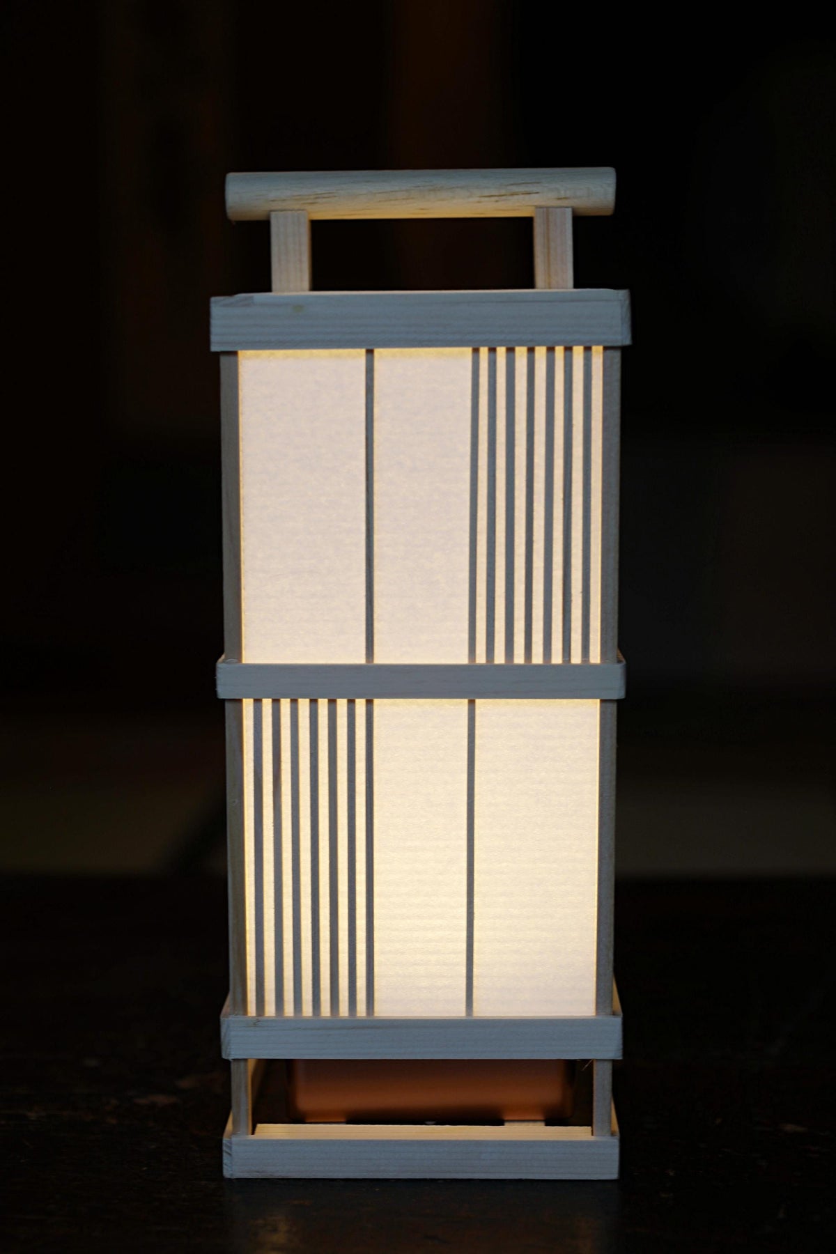 A white wooden lantern with a handle and a light inside, standing on a dark surface.