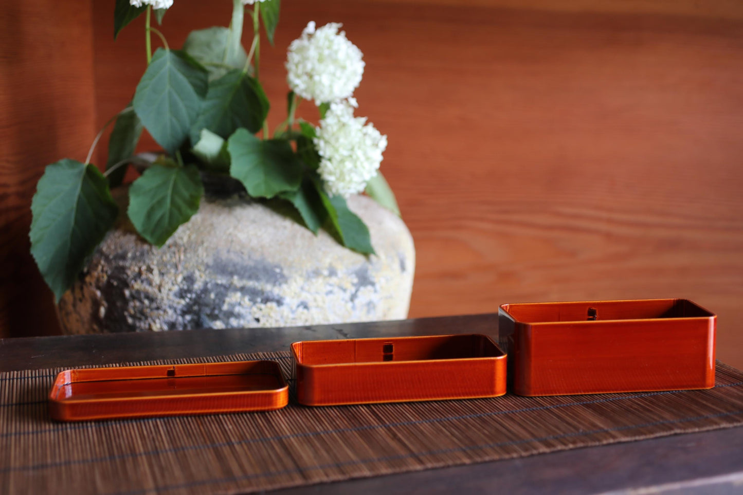 three red metal containers placed on a wooden surface, with a vase of white flowers in the background.