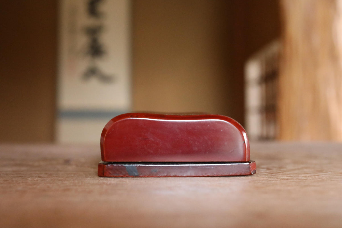 A red, curved object is sitting on a wooden surface, with a blurred background that includes a wall with Japanese characters.