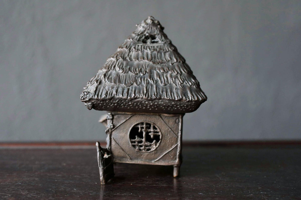 A small, intricately designed metal figurine of a house or hut, featuring a pointed roof and a circular window, sits on a dark surface.
