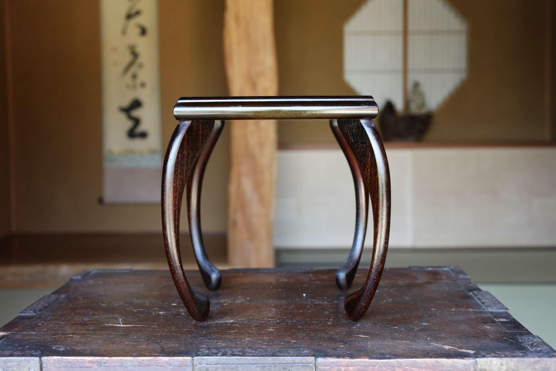 A wooden table with a curved top and legs, placed in a room with a window and a painting on the wall.