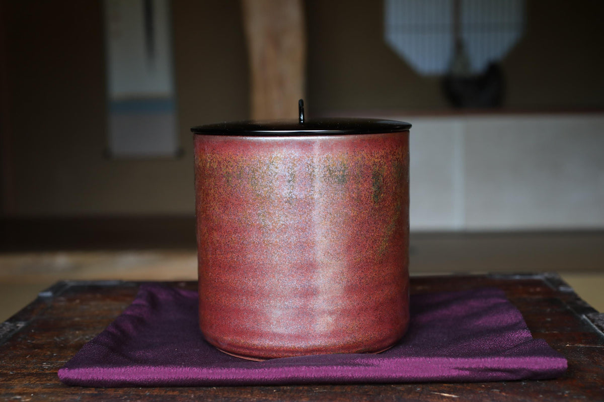 A red ceramic jar with a black lid sits on a purple cloth on a wooden surface.