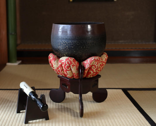 a large, dark bowl placed on a wooden stand, with a smaller bowl or container resting on top of it. The bowls are placed on a tatami mat, which is a traditional Japanese floor covering.