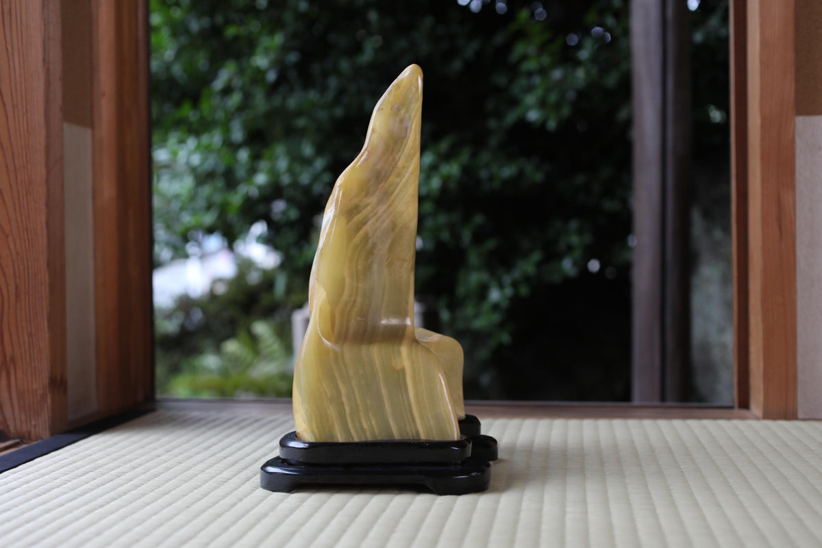 A yellow stone sculpture of a fish is displayed on a black stand in front of a window with a view of trees.