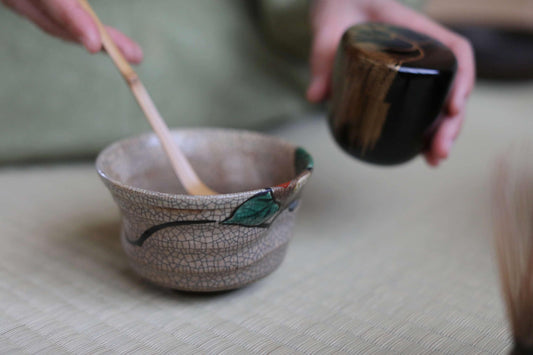 A person's hands are holding a small ceramic bowl with a green leaf design and a wooden spoon, suggesting they are preparing to serve tea or a similar beverage.