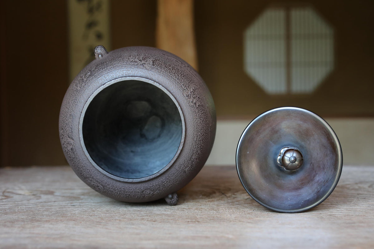 A ceramic teapot with a lid and a metal lid are placed on a wooden surface, with a traditional Japanese-style room in the background.