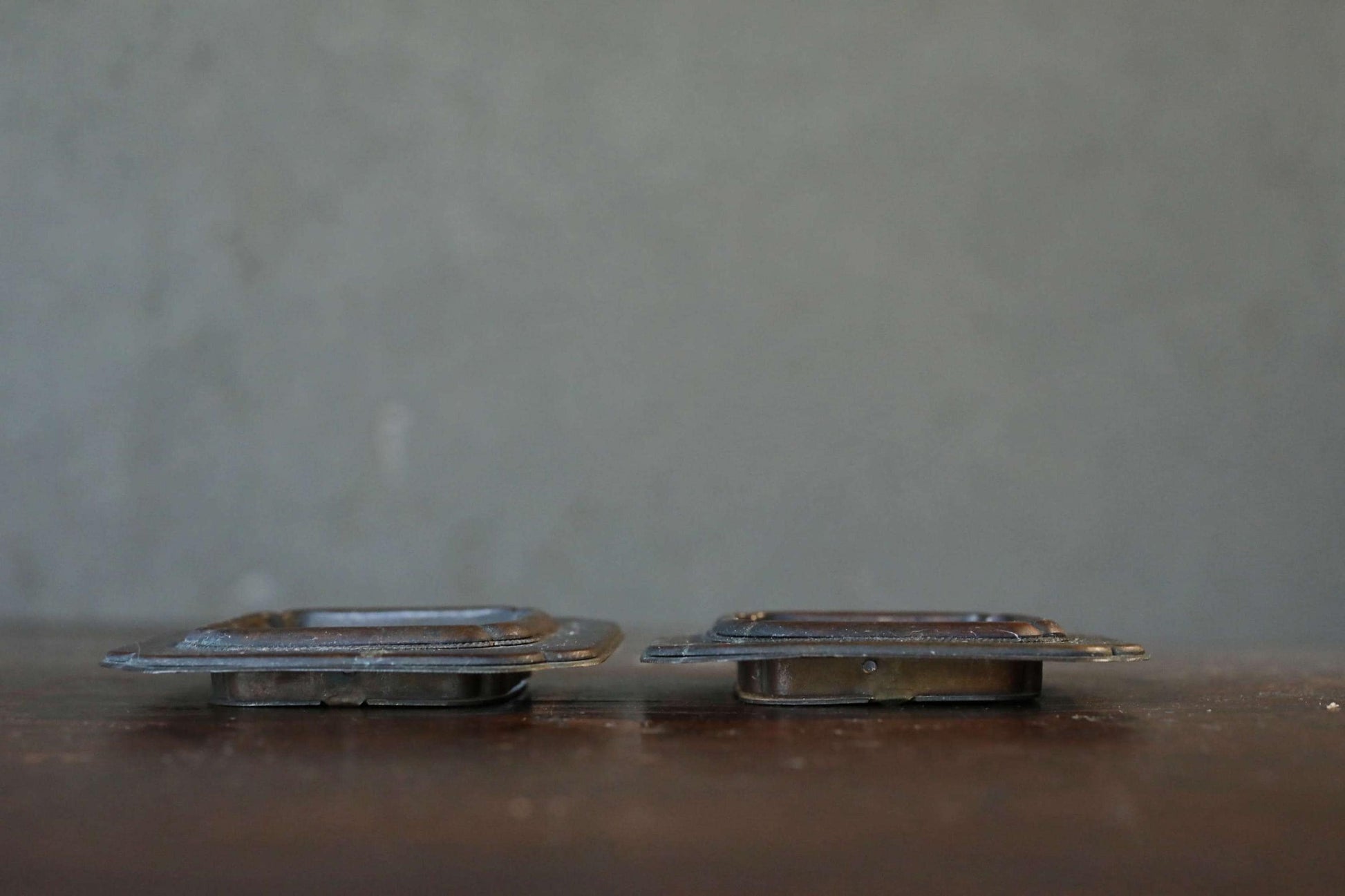 Two metal objects, possibly hinges or handles, are placed side by side on a wooden surface against a gray background.