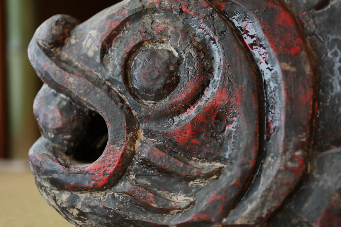 A close-up view of a weathered, red-painted wooden sculpture depicting a fish-like creature with a carved mouth and fins.
