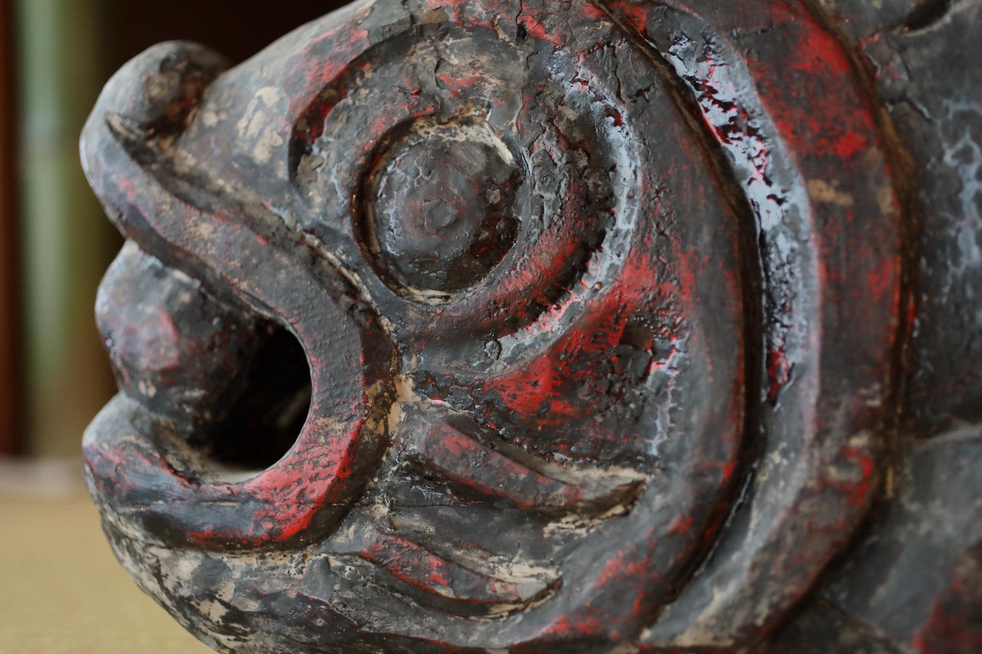 A close-up view of a weathered, red-painted wooden sculpture depicting a fish-like creature with a carved mouth and fins.