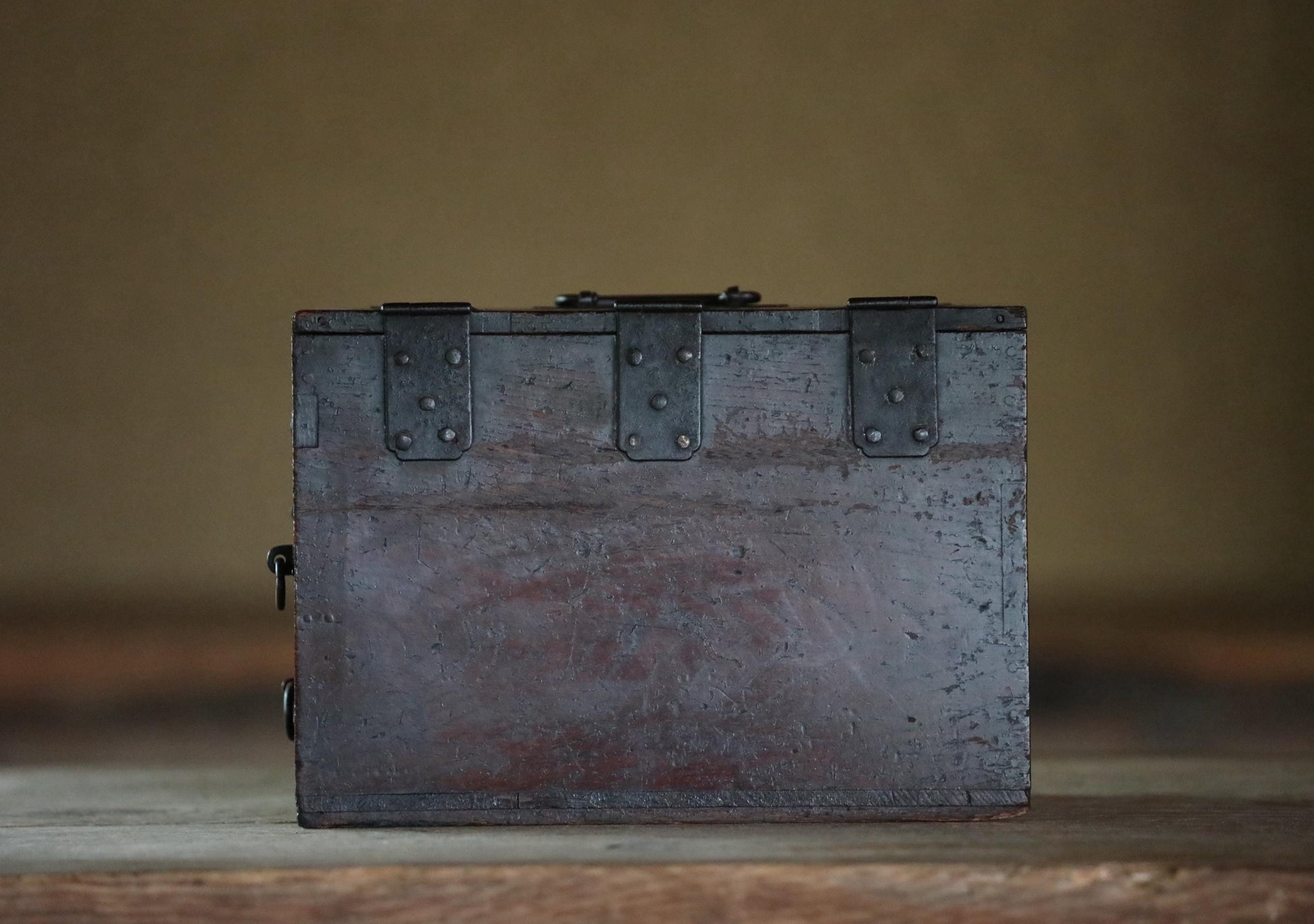 A weathered, wooden chest with metal clasps sits on a wooden surface against a plain background.