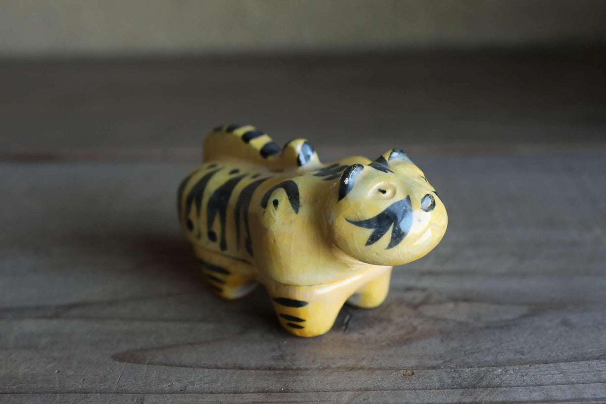 A ceramic figurine of a tiger-like animal with black stripes and a yellow body is sitting on a wooden surface.