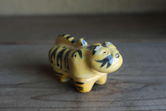 A ceramic figurine of a tiger-like animal with black stripes and a yellow body is sitting on a wooden surface.