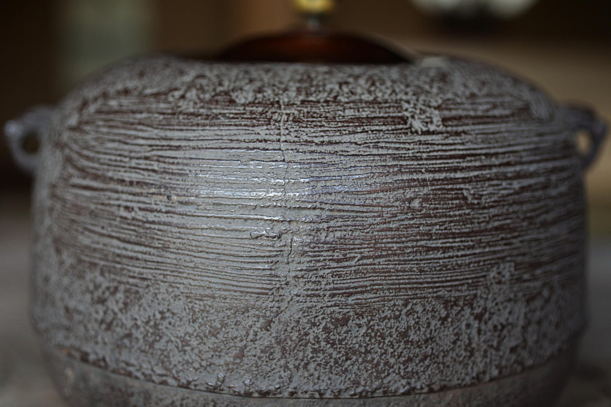 A close-up image of a textured, weathered pot or container with a wooden lid, possibly made of metal or ceramic material.