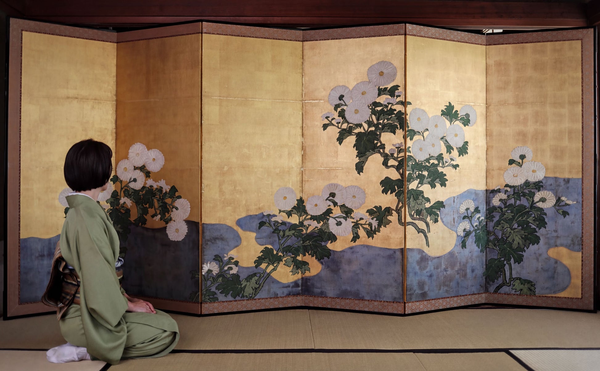 A person in a green kimono is kneeling in front of a large, intricately designed screen with floral and nature-inspired artwork.