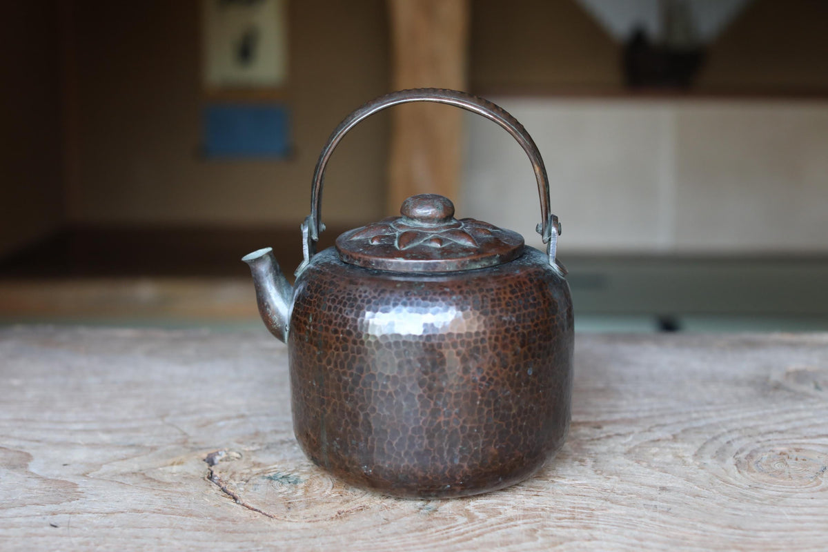 A shiny, copper-colored kettle with a handle and spout sits on a wooden surface.