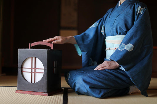 A person in a traditional Japanese kimono is kneeling on the floor, touching a small black lantern with a red handle.