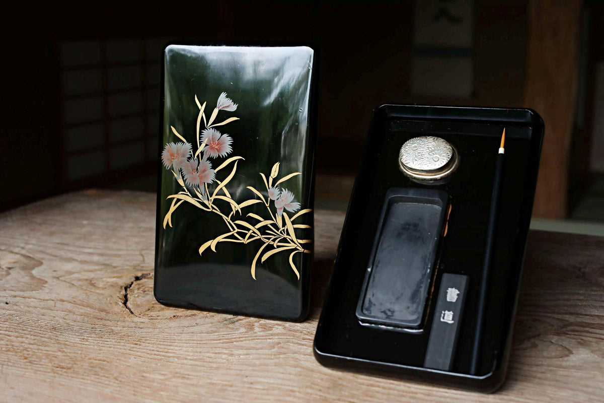 a black box with a floral design on it, placed on a wooden surface next to a black box with a silver object inside.