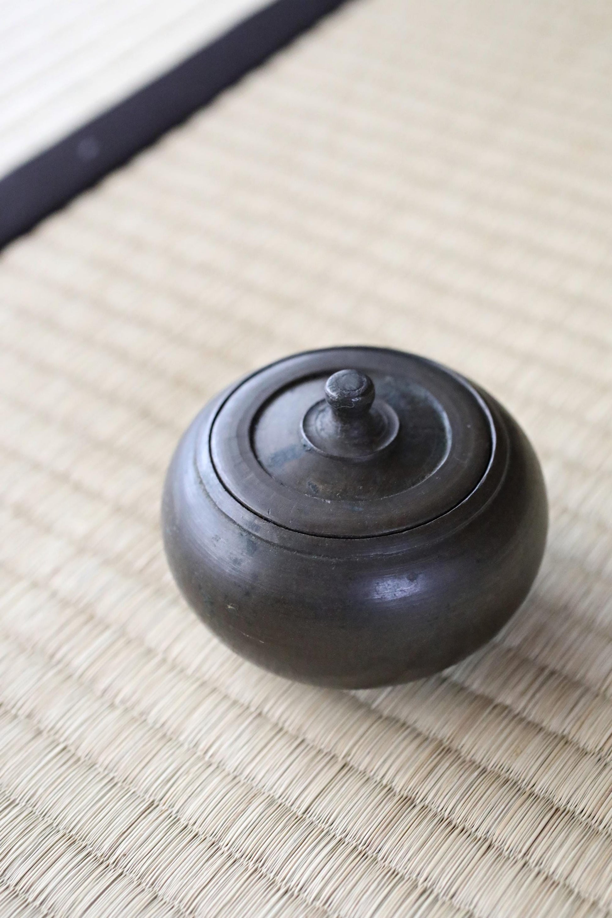 A black ceramic bowl with a lid sits on a woven mat.