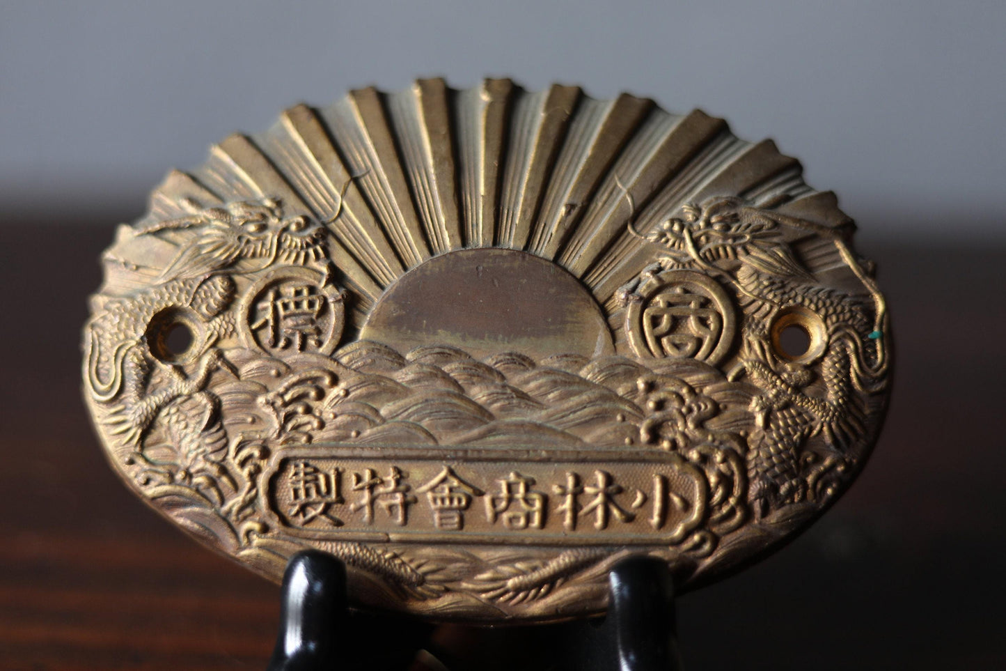 a decorative oval-shaped object with intricate designs and patterns, including a sun-like motif and two dragon-like figures. The object appears to be made of metal and is displayed on a stand.