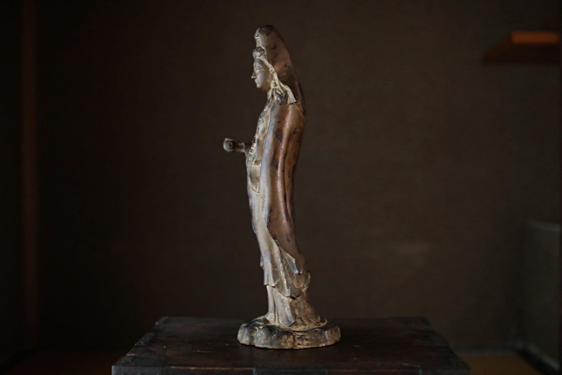A bronze statue of a woman with a long, flowing robe stands on a wooden surface against a dark background.