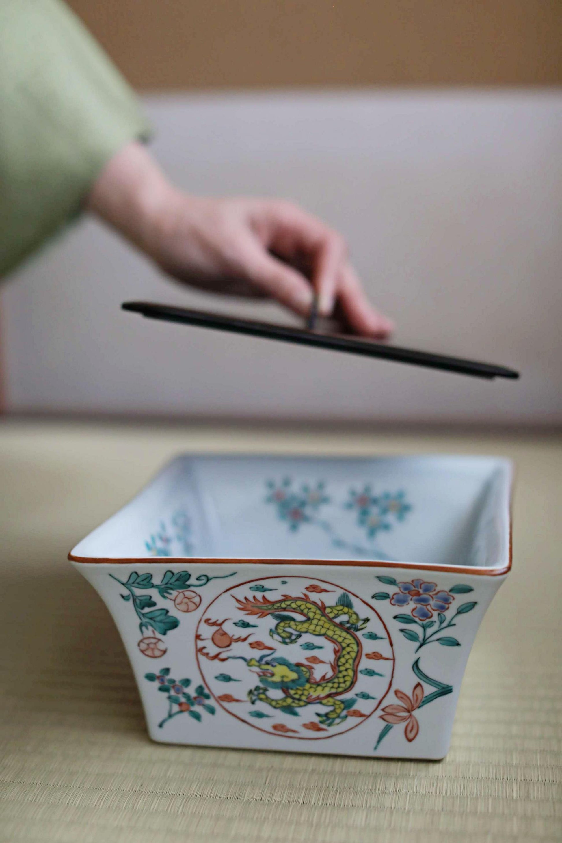 A hand is holding a black object above a white ceramic bowl with colorful floral designs.