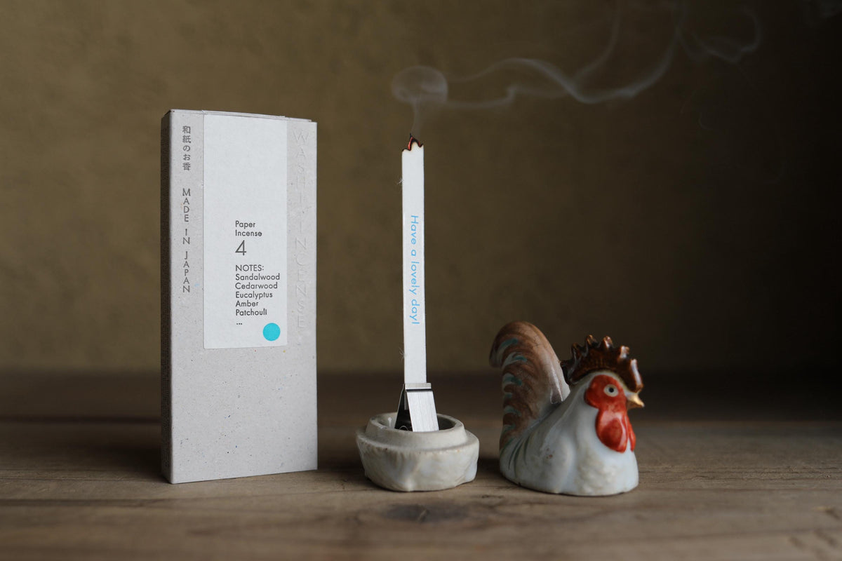 a white box with text on it, a ceramic rooster figurine, and a small incense holder on a wooden surface.