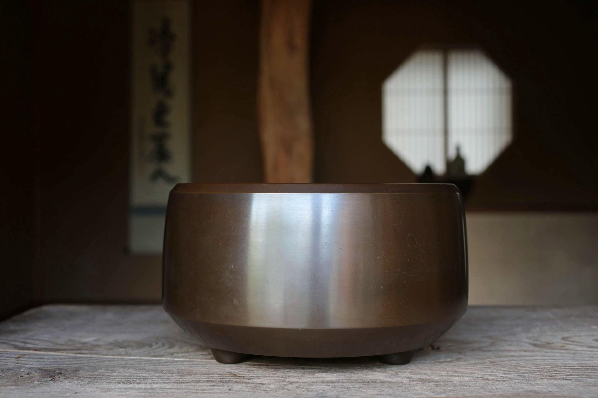 A shiny metal bowl sits on a wooden surface, with a window and a wooden beam visible in the background.