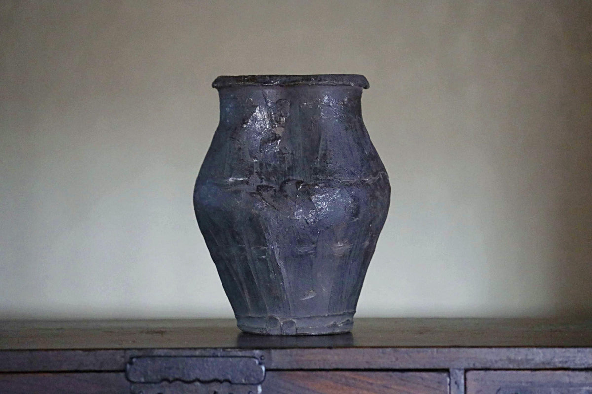 A dark-colored vase with a textured surface sits on a wooden surface against a plain background.