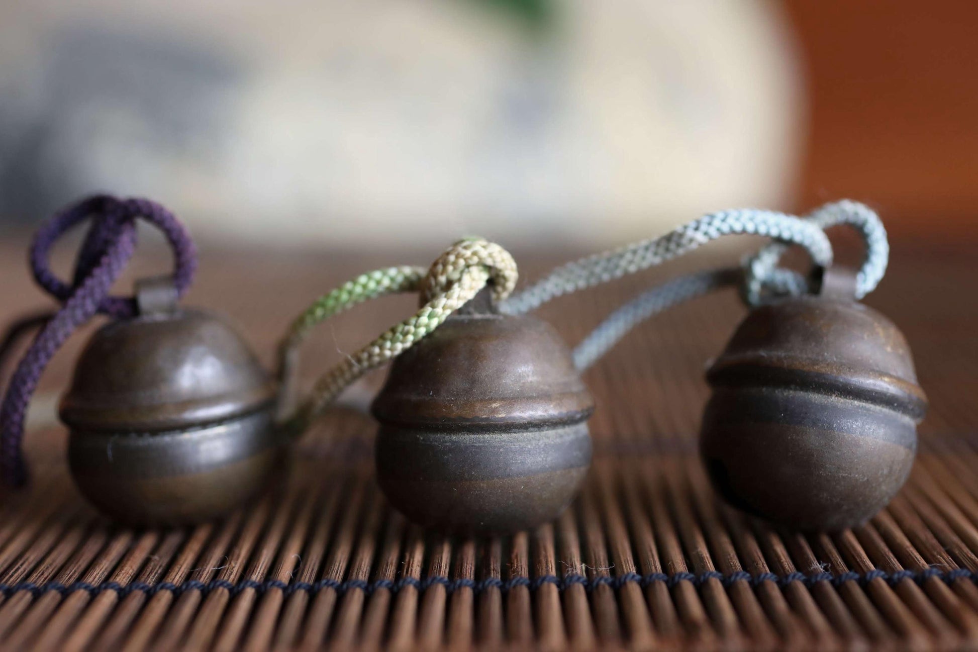 Three small, round brass bells with different colored strings attached to them, arranged on a bamboo mat.