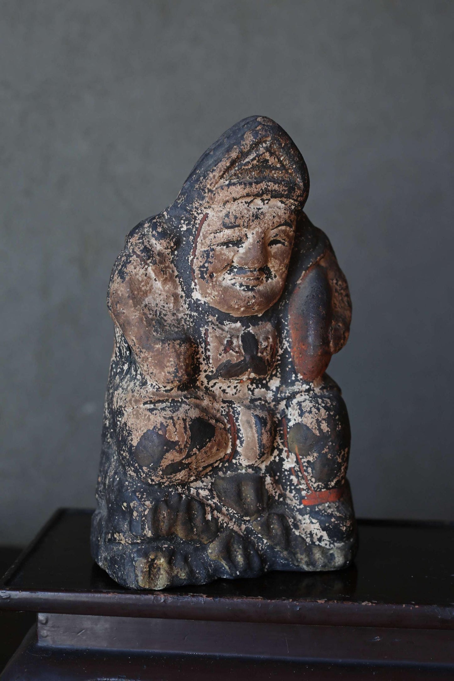 An old, weathered statue of a seated figure, possibly a deity or religious figure, displayed on a dark surface.