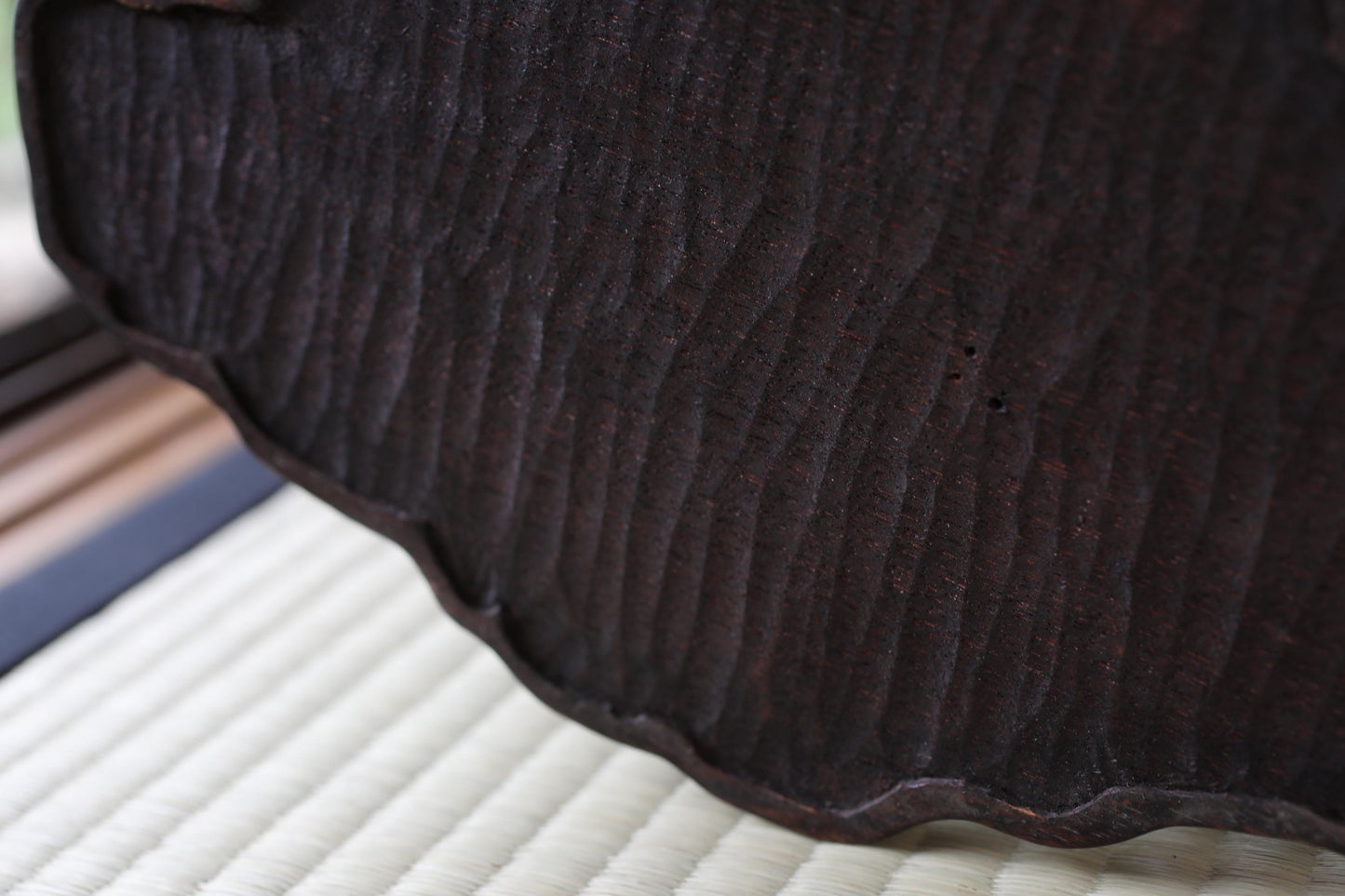 a close-up view of a dark, textured surface, possibly a piece of wood or metal, with a white woven mat or mat-like material in the background.