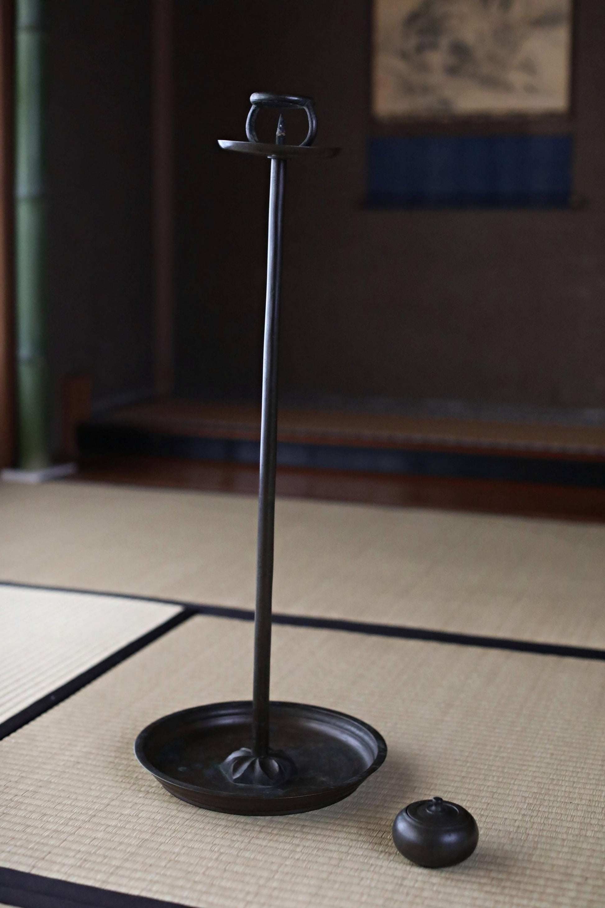 A black metal candle holder with a candle sits on a woven mat in a dimly lit room.