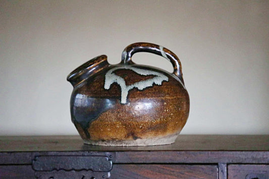 A brown and white ceramic pitcher with a handle and spout sits on a wooden surface.