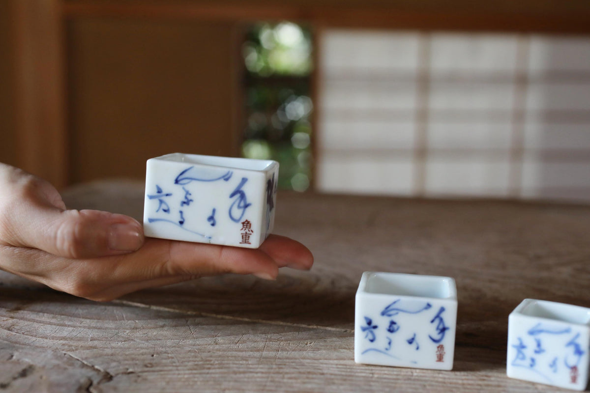 A hand holding a small, square ceramic container with blue designs on it, placed on a wooden surface.