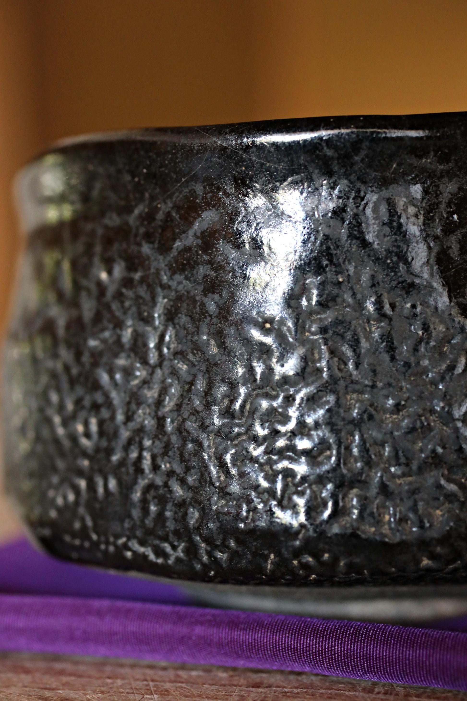 A close-up image of a black, textured ceramic bowl or vase placed on a purple surface.